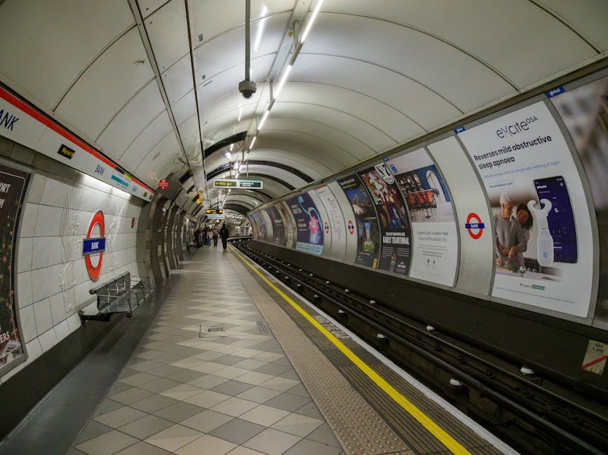 Interior view of a modern underground train station platform at High Barnet tube station in Barnet. The platform features a tiled floor with light and dark gray diamond patterns, illuminated by bright strip lighting on the curved ceiling. Along the walls are white ceramic tiles with the London Underground roundel sign and advertising posters displayed on the curved metallic walls. A yellow safety line runs parallel to the train tracks, and benches are visible along the wall. The scene shows a clean, well-maintained environment with no visible dust or debris, exemplifying professional surface and hygiene cleaning standards in a public transportation setting. Barnet Cleaner specializes in detailed sanitisation and deep cleaning for both residential and commercial spaces to maintain hygiene and cleanliness.